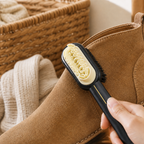 Black suede and nubuck shoe brush with cream grooved face and thin gold handle accent beside a tan boot