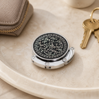 Ornate black and silver foldable purse hook stored compactly on a neutral vanity tray